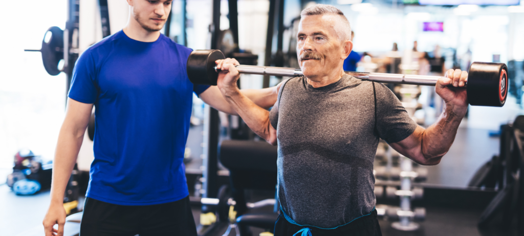 A young man in a blue shirt assisting an elderly man carrying a weightlifting equipment.